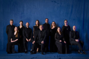 The Tallis Scholars wearing black formal attire pose in front of a blue fabric backdrop.