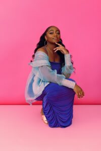 Samara Joy crouches in a pose in front of a pink wall wearing a vibrant blue dress and blouse.