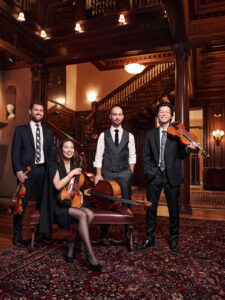 The Dover Quartet stand around a small couch in a dark wooden room with a staircase behind them, holding their instruments.