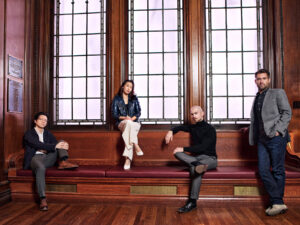 The Dover Quartet sit on a wooden bench below a wall of three large windows.