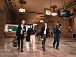 The Dover Quartet walk through a train station in casual clothing holding their instruments in travel cases.