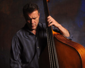 Edgar Meyer looking down while his hand rests on the strings of a large cello.