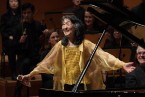 Mitsuko Uchida stands up to bow for the audience, wearing a flowy gold blouse.