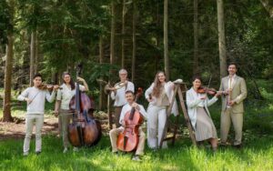 Members in the orchestra of the age of enlightenment pose with their instruments in a grassy field with the forest behind them.