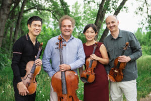 The Takacs Quartet smile with their instruments in a grassy field with trees behind them.