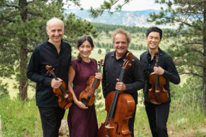 The Takacs Quartet smile with their instruments in a grassy field with trees and a view of the mountain behind them.