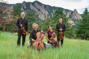 The Takacs Quartet smile with their instruments in a grassy field with trees and a tall mountain behind them.