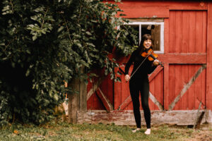 Tessa Lark smiles while standing in front of a red barn door with a large bush next to her, holding her violin.