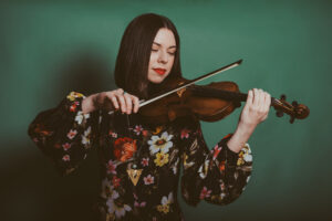 Tessa Lark wears a dark green floral dress and plays the violin in front of a green backdrop.
