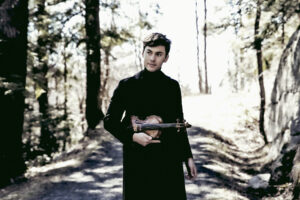 Benjamin Beilman wears a black trench coat and holds his violin in a forest clearing surrounded by tall trees.