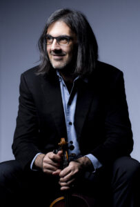 Leonidas Kavakos, with shoulder length grey hair, sits and holds his violin.
