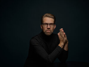 Dramatic photo of Lief Ove Andsnes wearing a black suit with his hands clasped in front of him.
