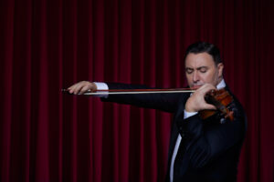Maxim Vengerov performs a violin solo in front of a red velvet curtain.