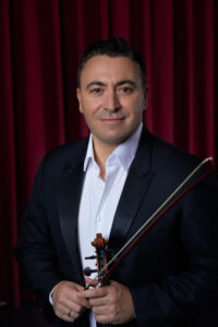 Maxim Vengerov wearing a formal suit and holding his violin in front of a red curtain.