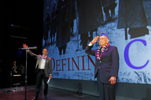 An elderly veteran salutes in front of a projection of Story Boldly's Defining Courage.