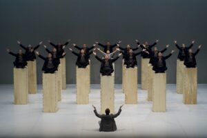 The monks of the Shaolin Temple sit atop tall wooden blocks onstage, looking upwards with their arms outstretched.