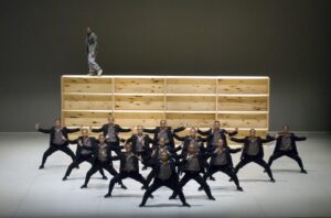 The monks of the Shaolin Temple stand in a triangle formation in a matching stance with wooden boxes behind them, one man standing atop.