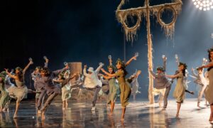 Dancers of the Joffrey Ballet wear flowy earth toned outfits onstage in matching poses, holding prop wineglasses in hand.