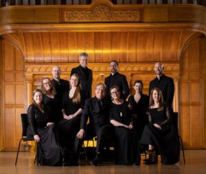 The Tallis Scholars smile happily at the camera while wearing black formal clothing, posing as a group in front of a gold wooden mantle.