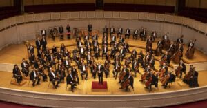 Riccardo Muti and the Chicago Symphony Orchestra hold their instruments and smile at the camera as they sit in formation onstage.