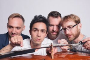 The four members of the JACK Quartet posing with serious expressions as if they're picking apart a cello and its bow.