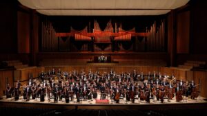 The Philharmonia Orchestra and Santtu-Matias Rouvali stand holding their instruments onstage in front of a large organ.
