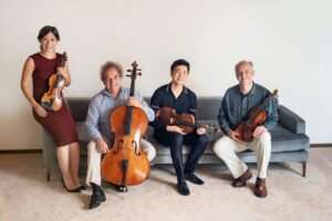 The Takács Quartet members, three men and one woman, sit smiling with their instruments in hand as they all sit on a long blue couch.