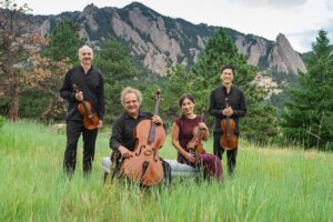 The Takács Quartet members, three men and one woman, smile with their instruments in hand in front of a lush forest background.