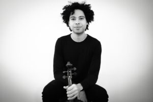 A black-and-white photo of Jordan Bak, a man with coily hair wearing all black, holding his viola and smiling softly at the camera.