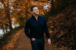 Aryeh Nussbaum, a young white man with short brown hair, wears a blue sweater smiling to his left in front a background of trees with orange leaves.