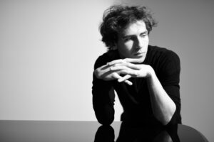 A black-and-white image of Alexandre Kantorow, a young French man, leaning against a piano with his hands together, looking away from the camera.