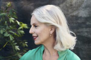 A left profile image of Anne Sofie von Otter, an older white woman with shoulder-length white hair wearing a green blouse, smiling brightly.