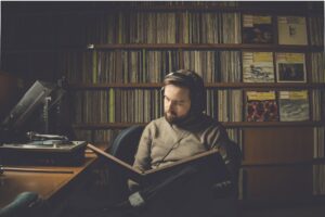 Daniil Trifonov, a young Russian man in a beige sweater, wears headphones connected to a record player as he reads in the middle of a vinyl record library