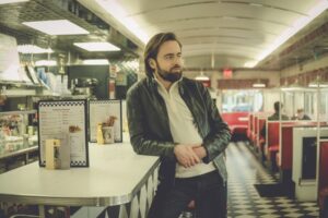 Daniil Trifonov, a young Russian man in a white quarter-zip and leather jacket, leans against the counter in a retro diner.