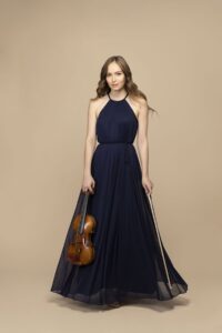 Geneva Lewis, a young woman with long curled brown hair wearing a navy blue dress, smiles as she poses with her violin in hand.