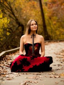 Geneva Lewis, a young woman with long brown hair wearing a red dress with black flowers, smiles to her side as she sits with her violin in her lap in front of a background of autumn leaves.