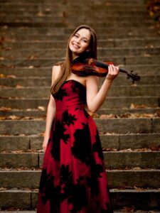 Geneva Lewis, a young woman with long brown hair wearing a red dress with black flowers, smiles as poses with her violin in front of a stone staircase.