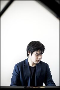 Nobuyuki Tsujii, a young Japanese man with dark hair wearing a navy blue suit, sits at a piano and looks down to his left in front of a white background.