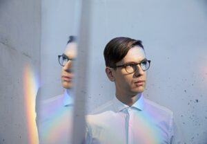 Vikingur Olafsson, an Icelandic man wearing glasses and a white button-up, stares to the left of the camera with a mirror reflecting his profile with soft rainbow lighting on him.