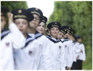 The boys of the Vienna Boys Choir smile as they wear matching uniforms and hats in front of a background of greenery.