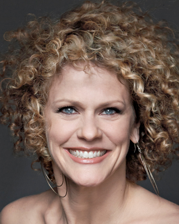 Artist Amber Martin, with a voluminous afro-style cut of blonde curly wears large hoop earrings and smiles, her eyeliner emphasizing her light blue eyes.
