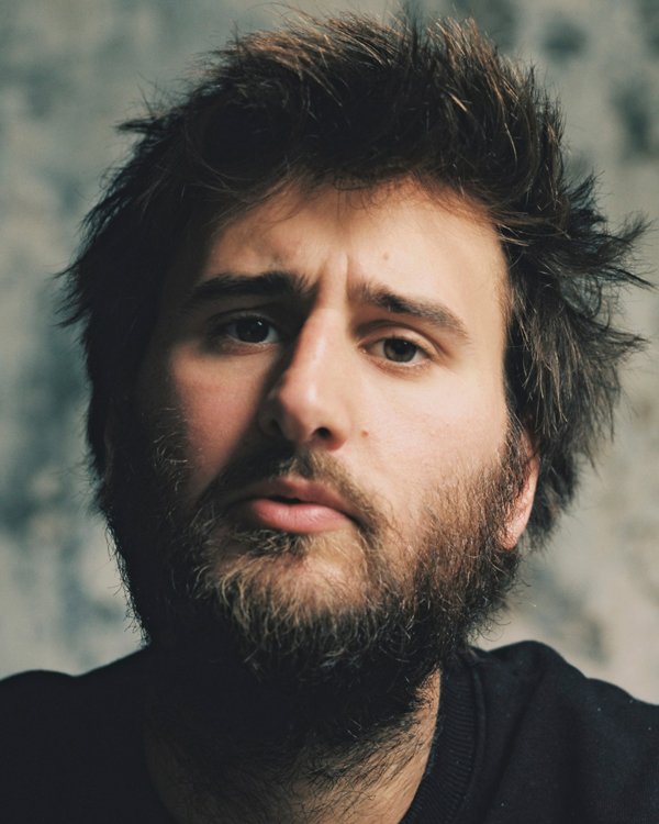 A sepia toned photograph of Artist Jean Rondeau, a young man with course brown hair and a beard.