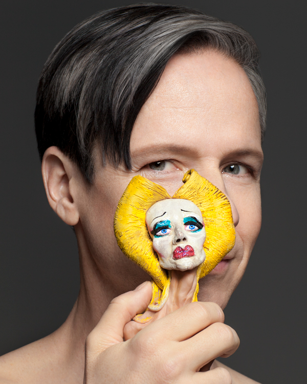 Artist John Cameron Mitchell holds a miniature abstract sculpture of a face with yellow hair, big red lips, and blue eye makeup in front of his own face.