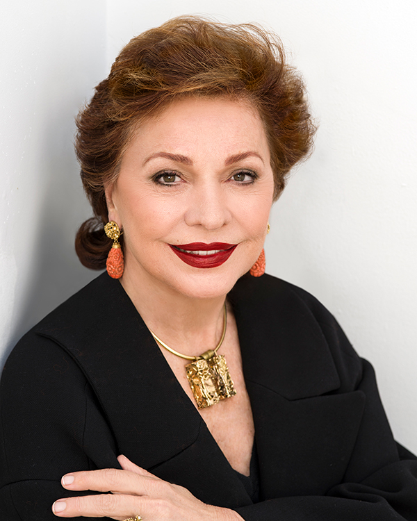 Maria Manetti Shrem smiles in a close up photo, wearing a black blouse, gold statement jewelry, and bold red lipstick.