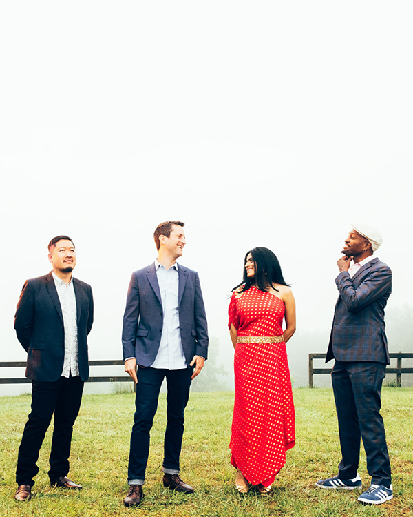 The members of the American Patchwork Quartet, three men wearing suits and one woman in a bright red dress, smile looking at each other