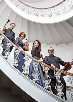 The Brentano String Quartet, two men and two women, hold their instruments and smile down at the camera from a white spiral staircase.