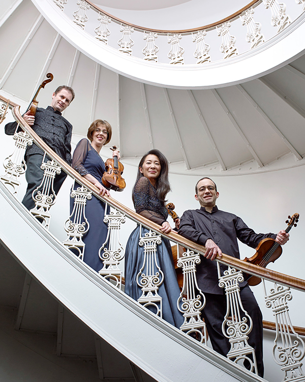 The Brentano String Quartet, two men and two women, hold their instruments and smile down at the camera from a white spiral staircase.