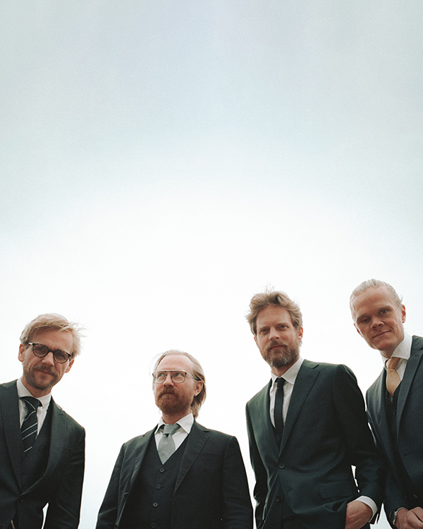 The four men of the Danish String stand next to each other, dressed in suits and ties, looking down at the camera below them.