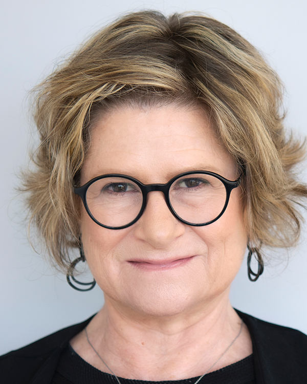 Dina Aldor, a white woman with short blonde hair and round black glasses, smiles into the camera