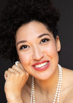 Julia Bullock, a woman with light olive skin and dark curly hair tied up, wearing a pearl necklace, smiles into the camera.